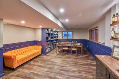 New basement home remodel with a bright orange accent couches and a small table in the back of the room. Off to the side is a built in storage and bookshelf