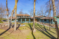 A modern two-story house with large windows sits among tall leafless trees and a grassy yard. There are outdoor seating areas, a fire pit, and climbing holds on one tree, with blue sky in the background.