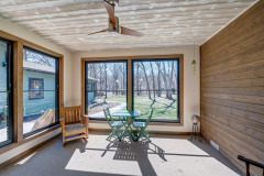 Bright sunroom addition remodel with large windows, a wooden ceiling fan, a small green table with two chairs, a wooden rocking chair, and a view of a yard with trees and a blue house outside.