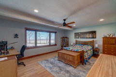 A spacious bedroom with a wooden bed, colorful quilt, desk with office chair, dresser, ceiling fan, large window with a scenic view, and hardwood floors. Artwork hangs above the bed.