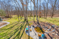 A spacious backyard with leafless trees, a stone patio with wicker furniture, and a small pond surrounded by rocks. Bright sunlight creates long tree shadows across the green lawn.