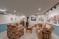 Cozy basement living room with beige sofas, a colorful armchair, a wood stove, and coffee tables, surrounded by framed photos and art on blue wainscoted walls under track lighting.