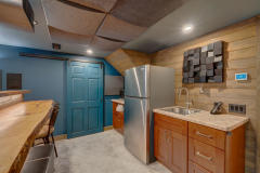 A modern kitchenette upgraded basement that features stainless steel appliances, wooden cabinets, a sink, a natural wood bar with stools, blue walls, a blue sliding barn door, and geometric wall art under recessed lighting.