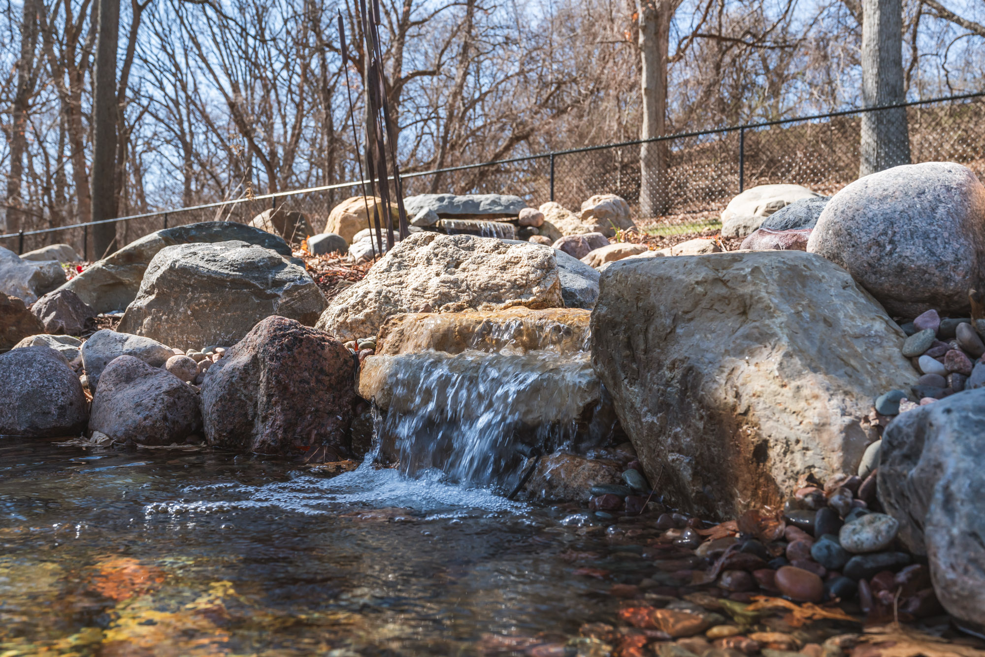 A small, clear stream cascades over rocks in a natural outdoor setting, surrounded by large stones, leaf litter, and bare trees. A chain-link fence runs along the background.