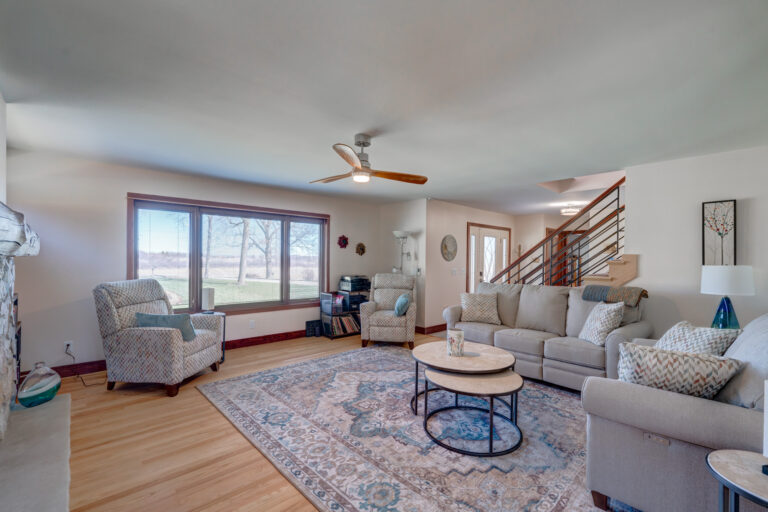 Bright living room with a large window, patterned rug, beige sectional sofa, two armchairs, round coffee tables, wood flooring, a ceiling fan, and a staircase with a modern railing in the background.