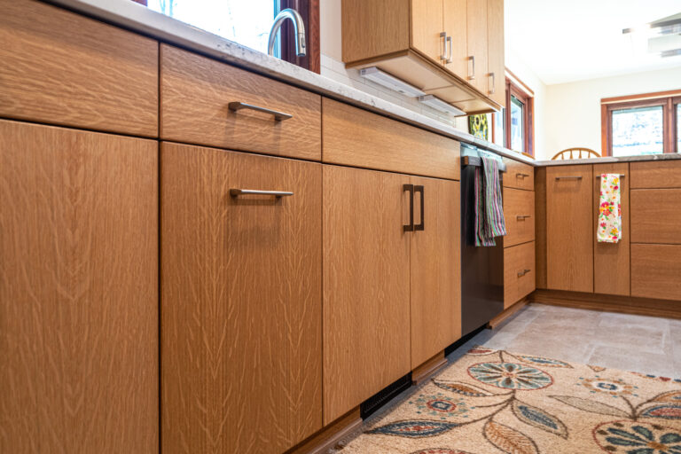 Modern kitchen with light wood cabinets, a marble countertop, stainless steel handles, a sink, colorful towels hanging, and a patterned rug on the floor. Natural light comes through large windows in the background.