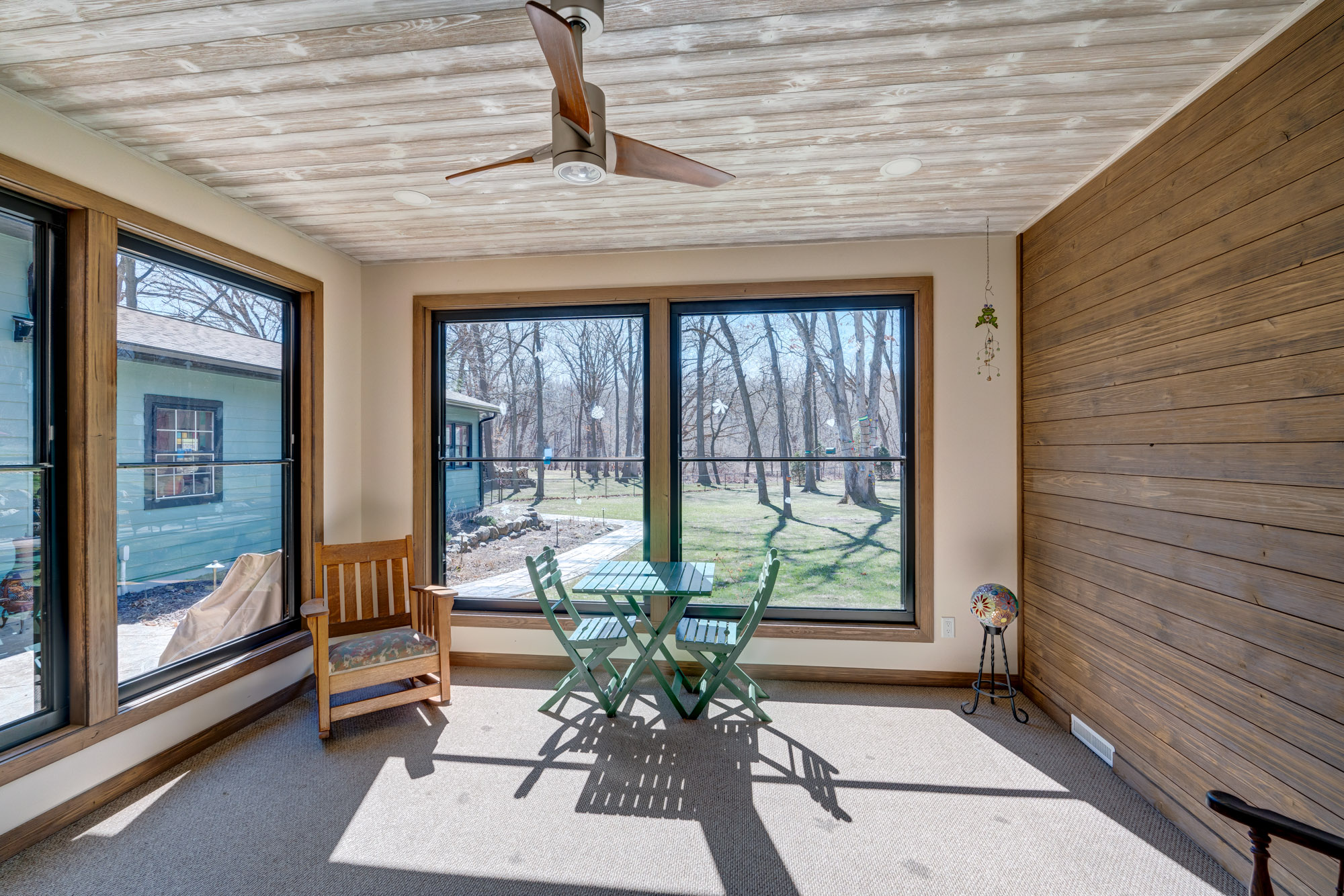 Bright sunroom with large windows, a wooden ceiling fan, a small green table with two chairs, a wooden rocking chair, and a view of a yard with trees and a blue house outside.