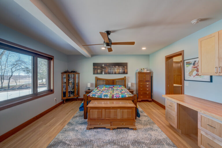 A cozy bedroom with a wooden bed, colorful quilt, wooden furniture, large window with trees outside, blue walls, ceiling fan, and decorative artwork above the bed.
