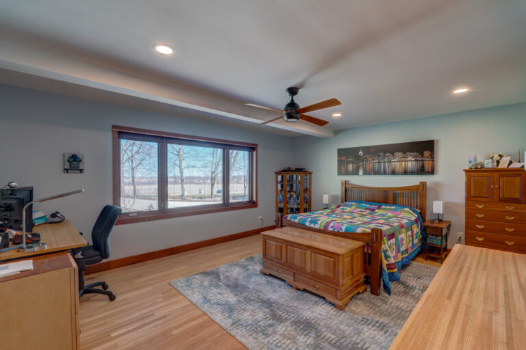 A spacious bedroom with a wooden bed, colorful quilt, desk with office chair, dresser, ceiling fan, large window with a scenic view, and hardwood floors. Artwork hangs above the bed.