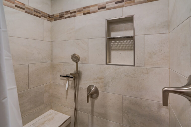Bathroom shower wall with accessibility seat and extra shower head. Tile is a tan marble with decorative rectangle strip. There is storage section built into the wall for users