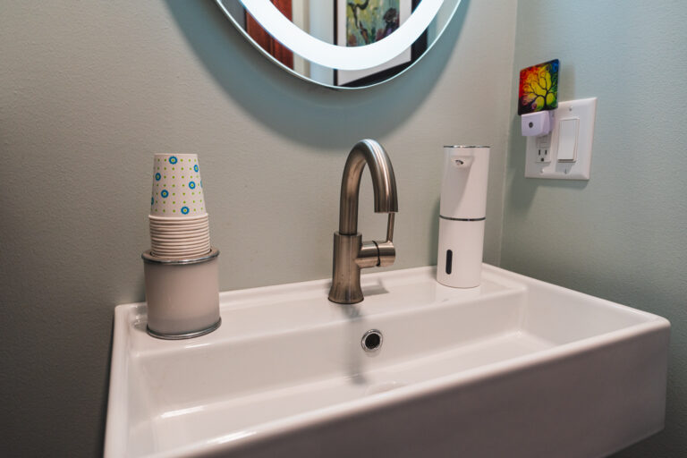 Modern white bathroom sink with silver fixture. Cups for mouthwash and a mini dispenser are on either side of the faucet