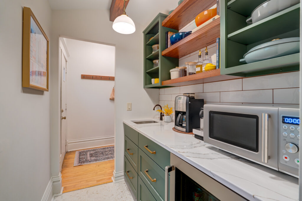 A small, modern kitchen with green cabinets, open wooden shelves holding colorful dishes, a coffee maker, a microwave, and a marble countertop. A hallway with wooden floors is visible through a doorway.