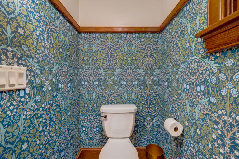 A white toilet is centered against a wall covered in blue and green floral wallpaper. A toilet paper holder with a roll is mounted on the right, and a small gold trash can sits on the floor. Wood trim lines the top of the wall.