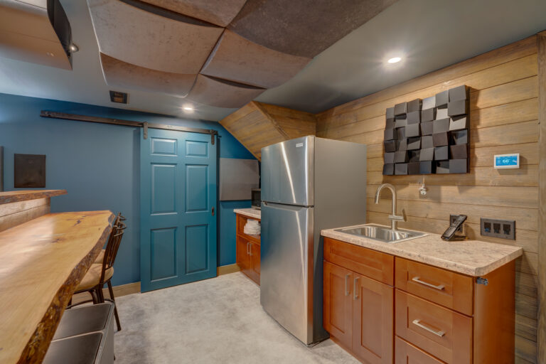 A modern kitchenette features wooden cabinets, a stainless steel refrigerator, a sink, a blue sliding barn door, unique ceiling panels, a wood-paneled wall, and a natural wood counter with bar stools.