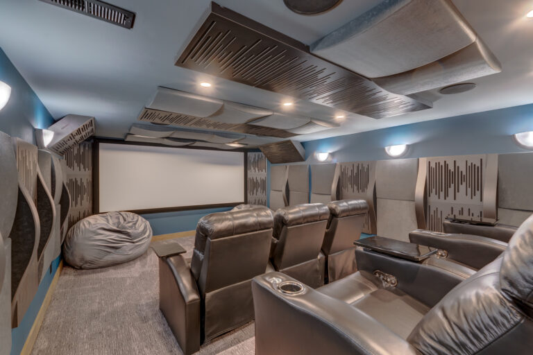 A modern home theater room with rows of black leather recliners, a large bean bag chair, acoustic panels on the walls and ceiling, and a big projection screen at the front under soft lighting.