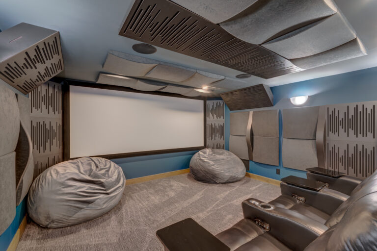 A modern home theater room with acoustic panels on the walls and ceiling, two large gray bean bag chairs on the floor, a row of black recliner seats, and a large projection screen at the front.