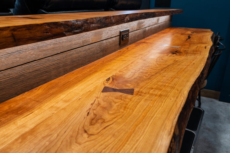 A close-up of a polished wooden bar top with a visible natural wood grain, featuring a butterfly joint detail and a built-in electrical outlet on the side panel. The background includes a blue wall.