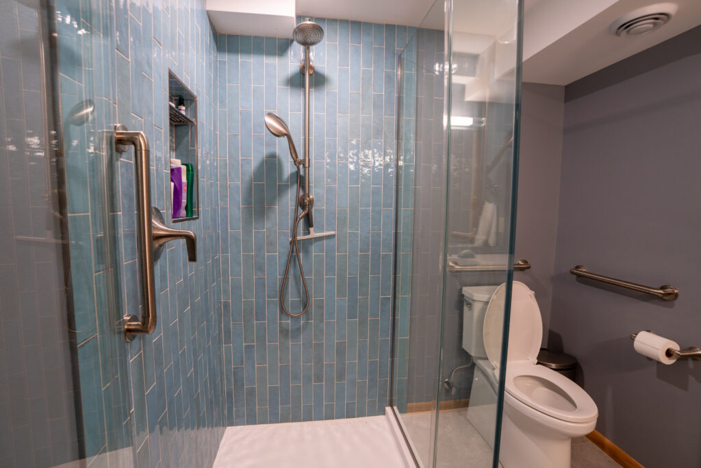 A modern bathroom with a glass-enclosed shower featuring blue subway tiles, a handheld showerhead, grab bars, and a built-in shelf. Next to the shower is a white toilet and a gray wall with a grab bar and toilet paper holder.