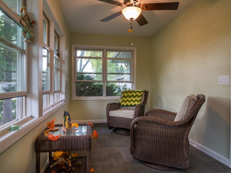 A cozy sunroom with wicker chairs, a small table decorated with autumn leaves, large windows letting in natural light, and a ceiling fan with a light above.