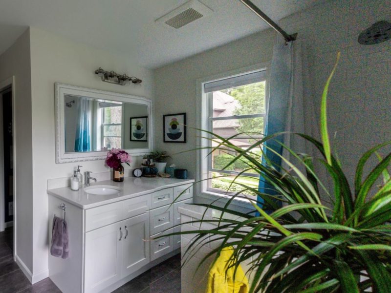 Bright bathroom with white cabinets and sink, wall mirror, potted plant in the foreground, a window letting in natural light, and a shower area with a blue curtain and yellow towel.